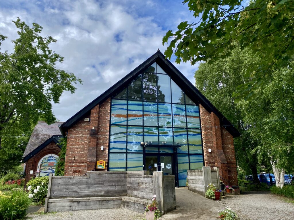 Chorlton Central Church - a contemporary building, low and wide. It's got a sloping roof that comes to a point in the centre, and almost the whole of the building is made from modern stained glass in flowing lines of white and blue, with a little yellow and orange and turquoise. It's surrounded by mature trees and is set in a small gravelled garden with roses in bloom. It has a ramp leading up to the double doors. 