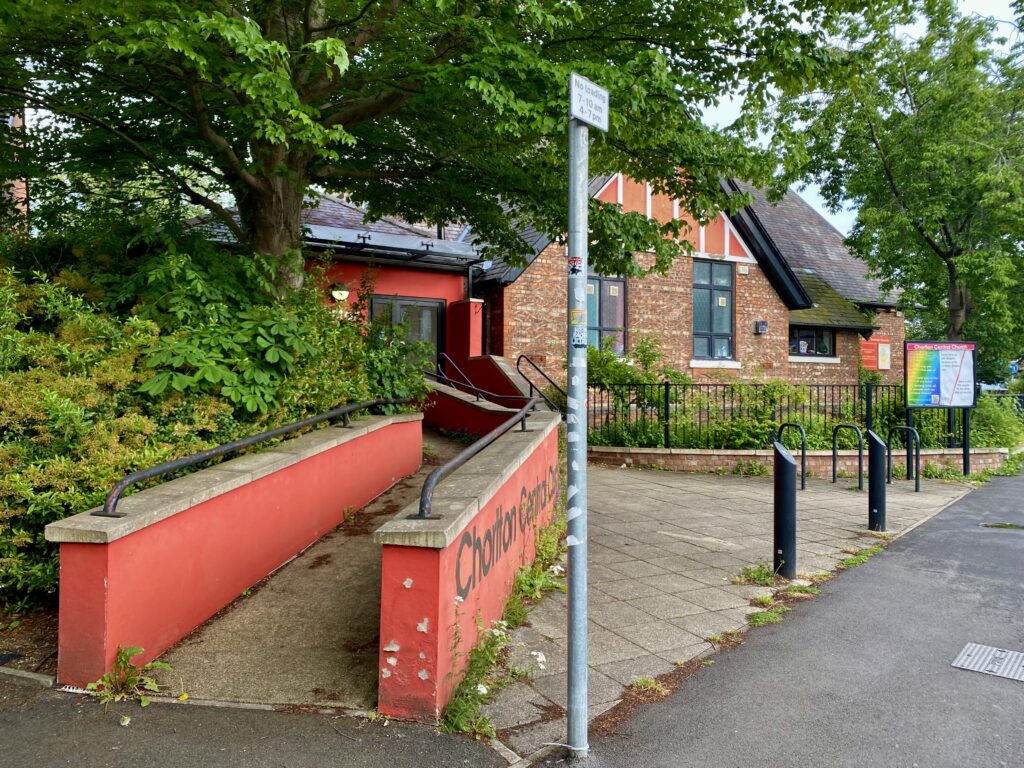 The side of the church has a wheelchair accessible entrance - a long cement ramp with rails on top of the low walls. The walls are painted a vivid orangey-red and one of them says Chorlton Central Church in large letters though it's hard to see in the picture. There's a street sign which says no loading between 7-10am and 4-7pm. 